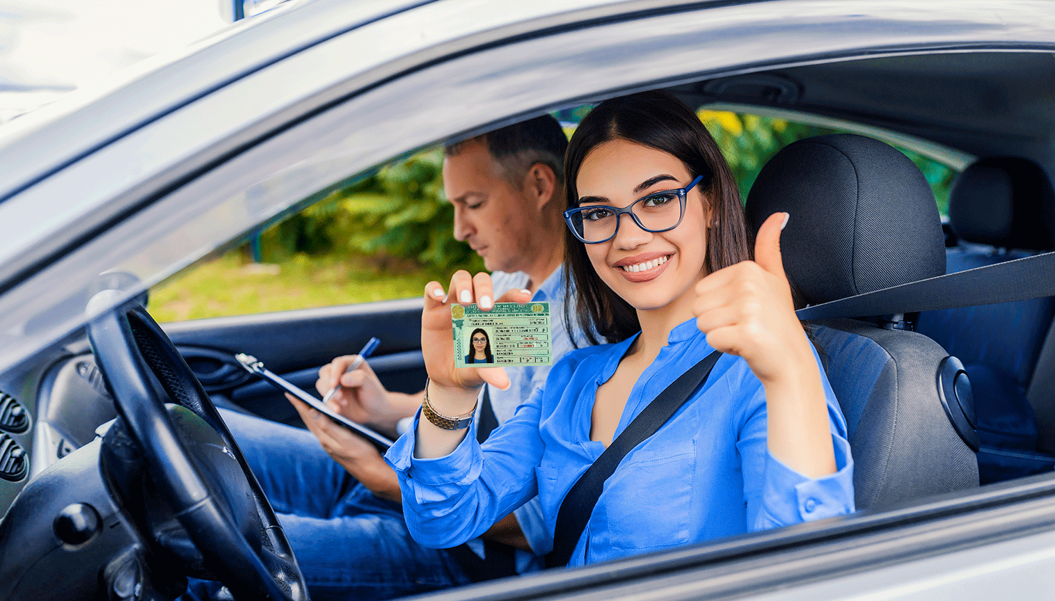 Na imagem, vemos ao fundo, dentro de um carro, um homem desfocado, já no primeiro plano vemos uma mulher sorrindo, usando o cinto de segurança, fazendo um sinal de positivo com uma mão e com a outra segurando uma carteira de habilitação (CNH).